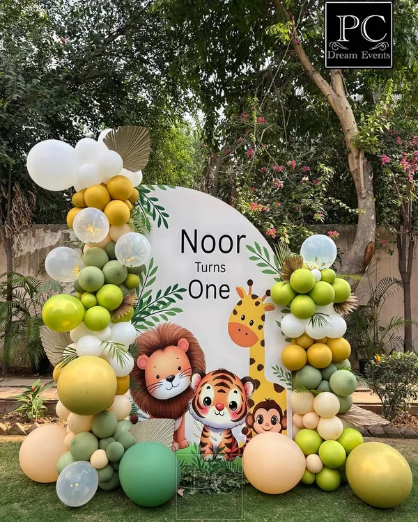 A custom circular backdrop for Noor's first birthday, featuring adorable safari animals like a lion, tiger, and giraffe. The theme is completed with organic balloon garlands in shades of green, yellow, and white.