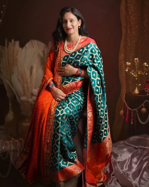 A full-length portrait of the mother-to-be in a stunning teal and orange saree. The studio setup includes pampas grass and traditional decor to complement the ethnic elegance of the attire.