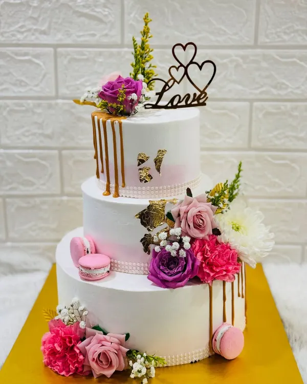 A magnificent three-tiered anniversary cake adorned with a cascade of fresh flowers, macarons, and a golden drip effect. A true centerpiece for a grand celebration of love.