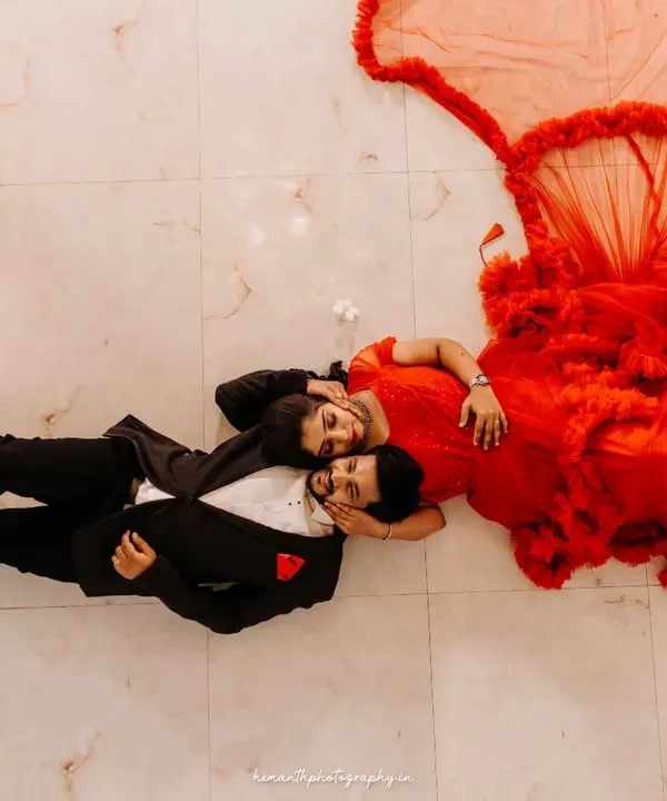 An artistic top-down shot of a couple lying on a marble floor. The bride's dramatic red gown flows out, creating a stunning, high-fashion portrait.