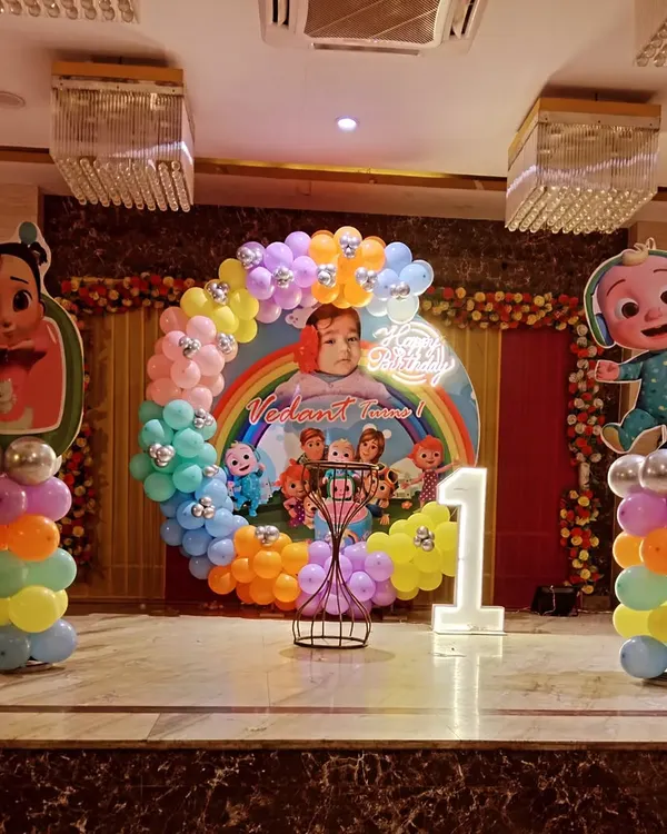 A full-stage Cocomelon theme for a first birthday party. This setup includes a large circular backdrop with characters, rainbow balloon arch, light-up number '1', and character standees on balloon pillars.