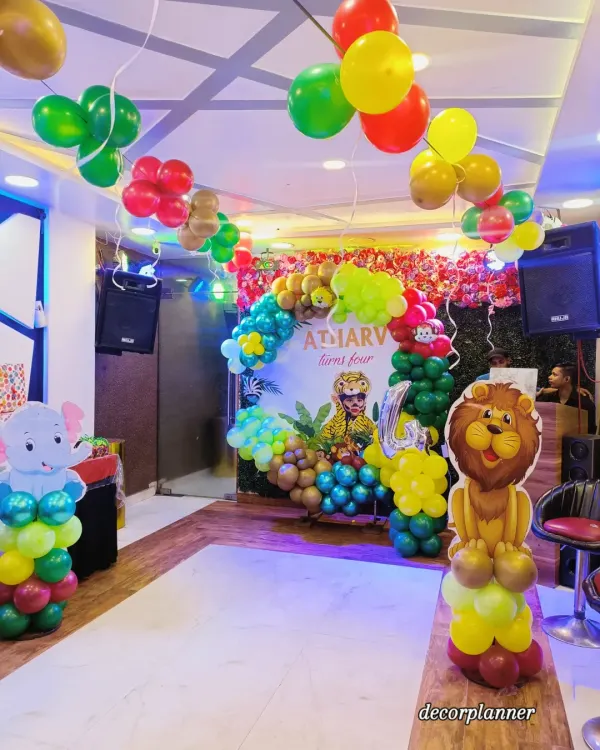 A full view of a fantastic jungle-themed party for a fourth birthday. The setup includes a large balloon arch over a personalized backdrop, cute animal standees like a lion and elephant, and balloon bunches hanging from the ceiling to create a complete jungle canopy effect.