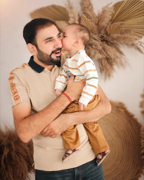 A father receiving a sweet kiss from his son during their boho-themed family photoshoot. The natural, earthy backdrop with pampas grass complements the warmth of this candid moment.