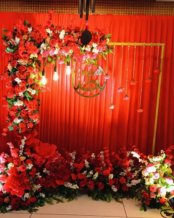 A vibrant and traditional backdrop for a pre-wedding ceremony. This setup features a rich red drape, a lush arch of red and white roses, and a custom gold acrylic sign with the couple's names.