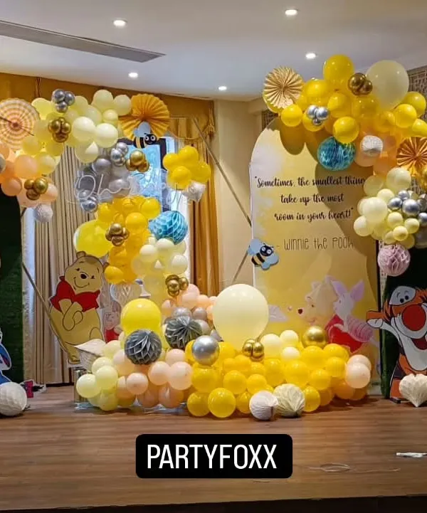A charming Winnie the Pooh themed baby shower. The setup includes character cutouts, a custom backdrop with a quote, and a flowing yellow and white balloon arrangement.