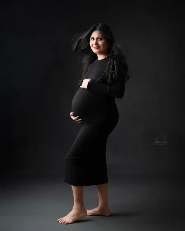 A powerful and modern solo portrait. The mom-to-be stands confidently in a simple black bodycon dress, with her hair blowing for a dynamic effect.