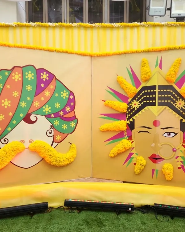 A funky and artistic photo booth for a Haldi ceremony. This backdrop features quirky illustrations of a bride and groom, with 3D elements made from fresh genda phool (marigolds).