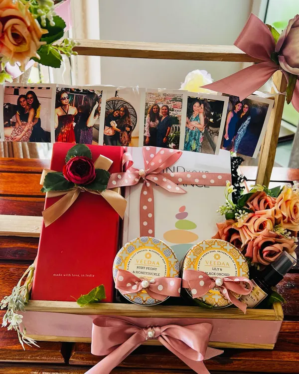 A close-up of a personalized hamper created for a best friend's wedding. The highlight is a string of cherished photographs, complemented by VILASA luxury wellness products and a gift box, all arranged in a beautiful wooden crate with pink floral details.