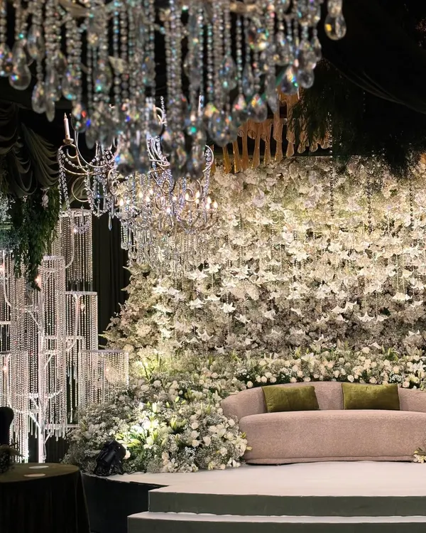 A full view of the enchanted forest stage, where a massive wall of white flowers serves as the backdrop. Crystal chandeliers and candle installations add layers of light and luxury to the magical setting.