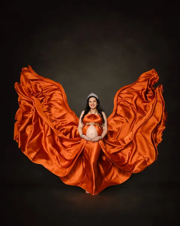 A breathtaking solo portrait using fabric tossing to create a dynamic, wing-like effect with a fiery orange satin gown. This technique adds movement and drama, making for a powerful and artistic maternity photo against a simple dark background.