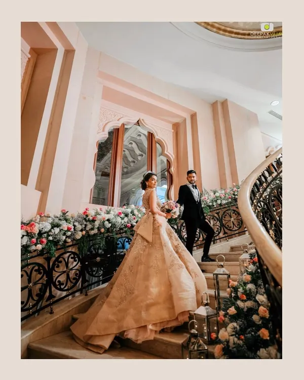 A fairytale moment on the grand staircase of The Leela Palace Bengaluru. The bride's magnificent gown flows behind her, perfectly framed by the floral-adorned railings we designed for their elegant reception.