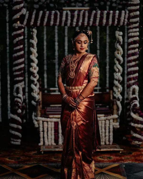 A full view of the traditional Tamil Brahmin bridal look, featuring the 9-yard Madisar saree. The rich maroon silk and intricate jewelry come together for a look of pure elegance in front of the Oonjal.
