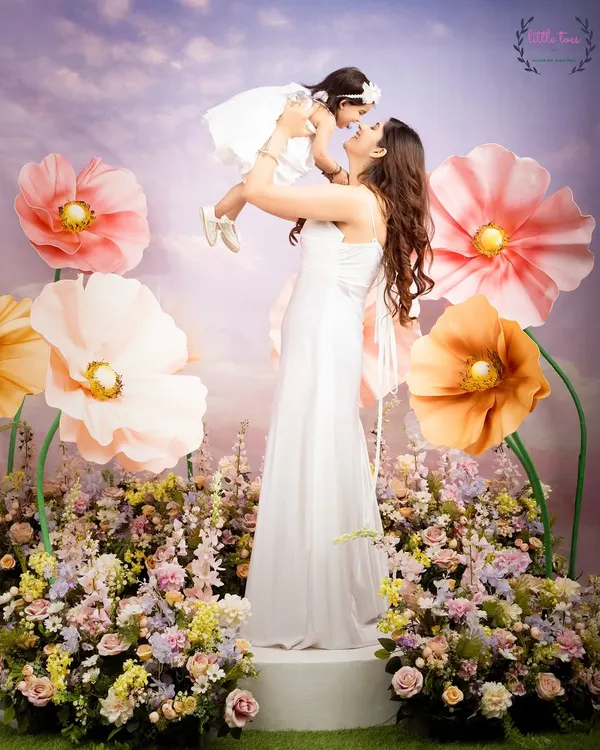 A mother joyfully lifts her daughter in the air, both dressed in white, standing in a field of oversized, whimsical flowers. This image is a celebration of the pure, uplifting joy of motherhood.