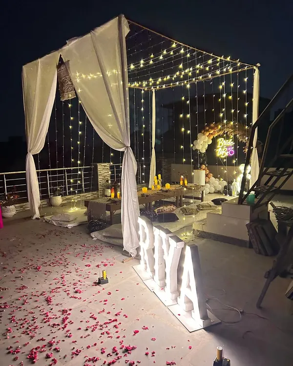 A wide view of a 'Bride to Be' celebration on a terrace, featuring a draped cabana illuminated by fairy lights. The ground is scattered with rose petals, leading to an intimate low-seating dinner area.