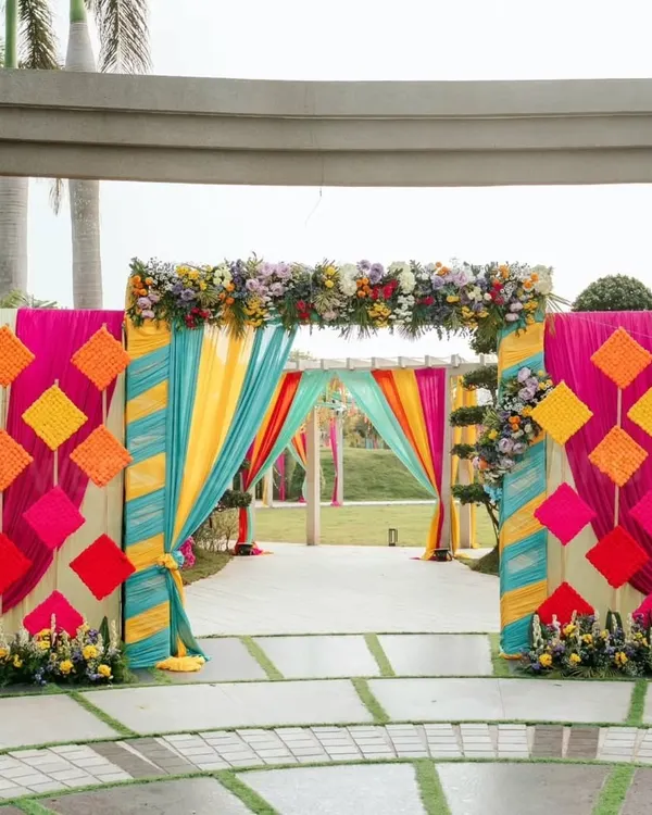 A grand entrance that screams celebration. The mix of colorful drapes in yellow, teal, and pink with geometric hangings and floral arrangements sets a festive tone from the moment guests arrive for the mehendi.
