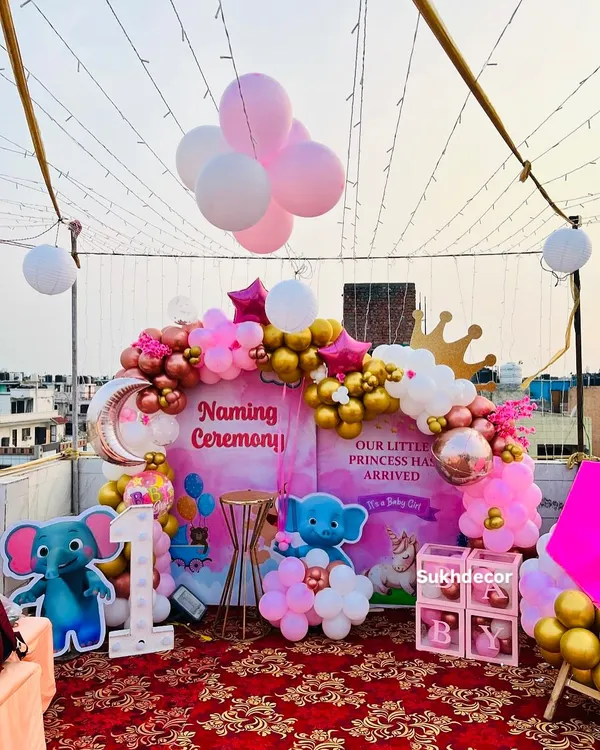A grand and festive rooftop setup for a princess-themed naming ceremony. The decor is built around a storybook-style backdrop and features a balloon arch in pink, white, and gold, character cutouts, and a light-up number '1'.