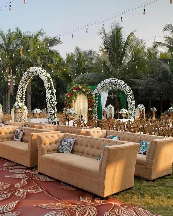 An outdoor daytime reception setup with elegant lounge seating. The white floral arches and lush palm trees create a beautiful, relaxed atmosphere for guests to mingle and celebrate.