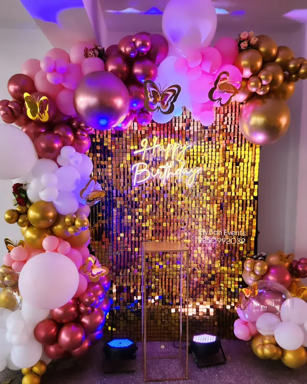 A vibrant birthday backdrop featuring a gold sequin shimmer wall. The wall is framed by an elaborate balloon garland in shades of pink, white, and gold, and is finished with a 'Happy Birthday' neon sign.