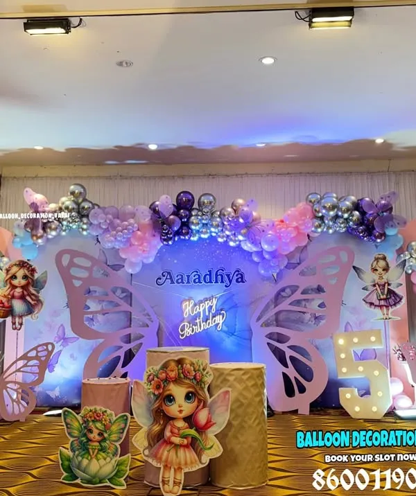 A grand butterfly and fairy theme for a 5th birthday, set up in a banquet hall. This decoration features large, illuminated butterfly wing props, custom character cutouts, and a beautiful balloon arch in shades of pink and purple over a themed backdrop.