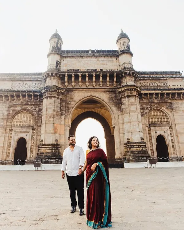 A grand, wide-angle shot featuring the couple at the Gateway of India. This image is a classic Mumbai pre-wedding photograph, celebrating their love story in the heart of the city.