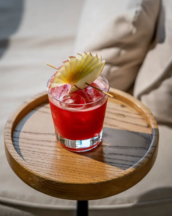A perfect lifestyle shot of a vibrant red cocktail. The natural sunlight, wooden tray, and soft focus background create a relaxed, upscale daytime drinking vibe. This is ideal for a hotel bar or a chic cafe's social media.