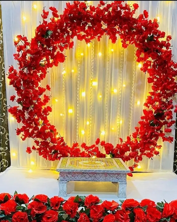A beautiful circular frame of red flowers set against a white curtain with pearl strings and fairy lights. This setup is ready for your idol, providing a vibrant and glowing backdrop.
