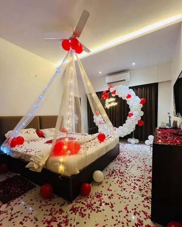 A complete room transformation featuring a circular balloon arch with a neon sign, a fairy light canopy, and a floor covered in rose petals. This red and white theme is a popular choice for romantic anniversary and birthday celebrations at home.
