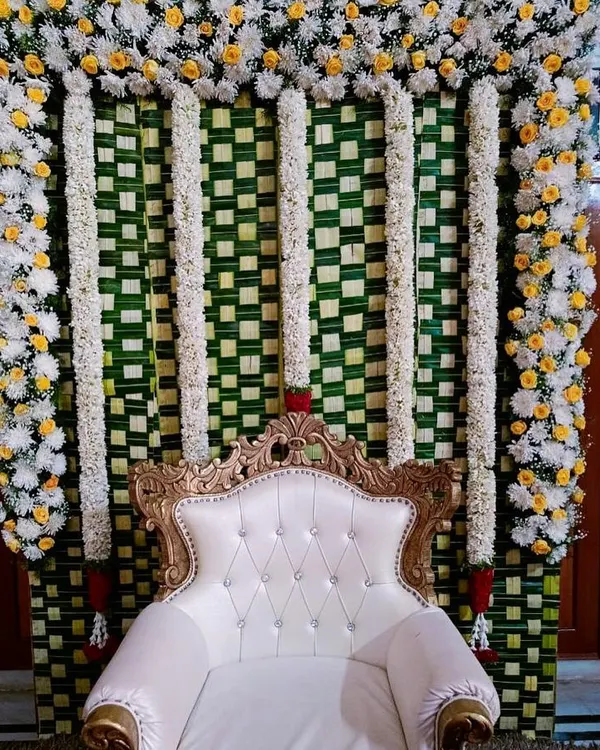 A complete traditional seemantha stage. The backdrop is made of woven coconut leaves, framed with white and yellow flowers, and features an elegant, tufted chair for the main ceremony.