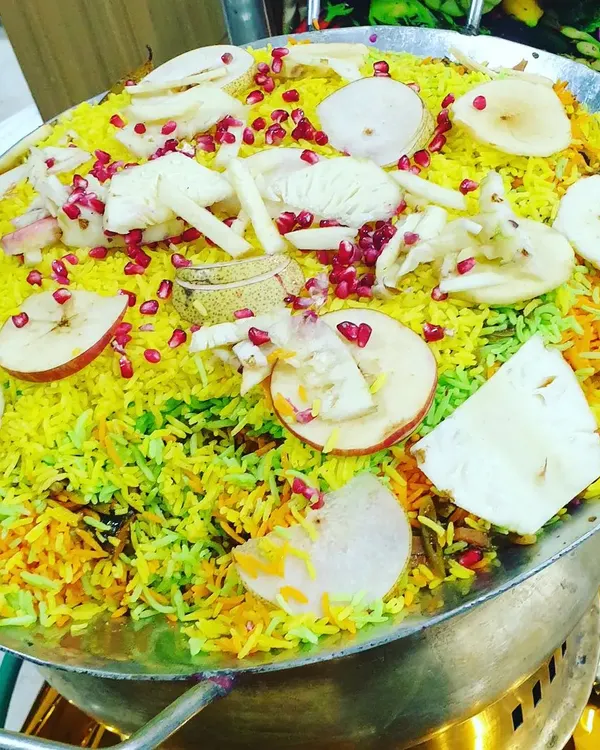 A large pan of colorful Kashmiri Pulao, a fragrant rice dish garnished with fresh fruits like apple and pomegranate. This is a visually appealing and delicious main course option.
