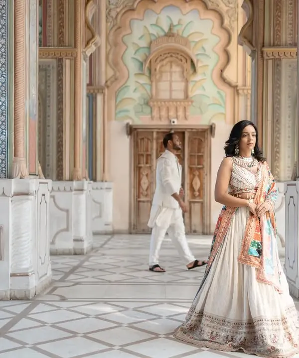 A portrait of modern royalty in a Jaipur palace. The bride stands poised in an elegant lehenga, while her partner walks in the background, creating a sense of depth and story within the frame. This is a perfect example of a destination heritage shoot.