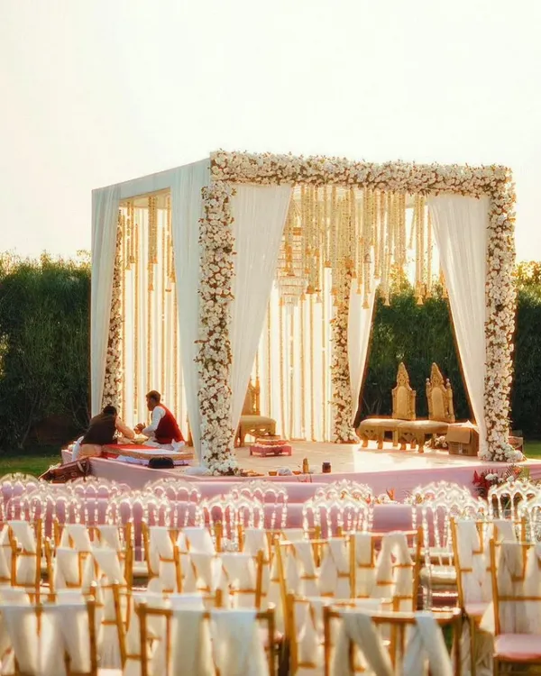 An outdoor wedding mandap with a clean, modern design. The structure is adorned with white flowers and flowing drapes, creating an elegant and timeless look for a daytime ceremony.