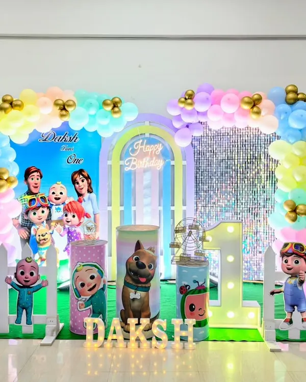 A bright and colorful Cocomelon birthday party setup. This decoration includes multiple character cutouts, a pastel rainbow balloon arch, and a large light-up number '1'.