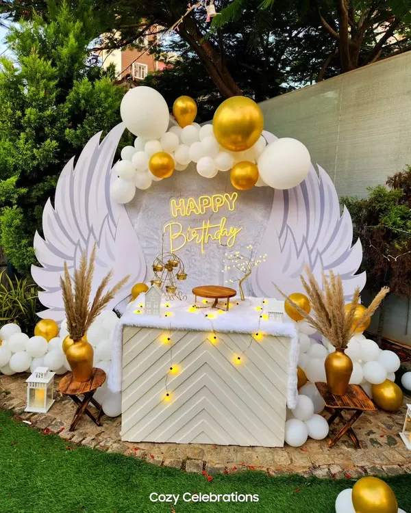 An ethereal angel wings backdrop for a birthday celebration. This heavenly setup in white and gold includes a custom-designed table, pampas grass, and a "Happy Birthday" sign, creating a truly unique photo opportunity.
