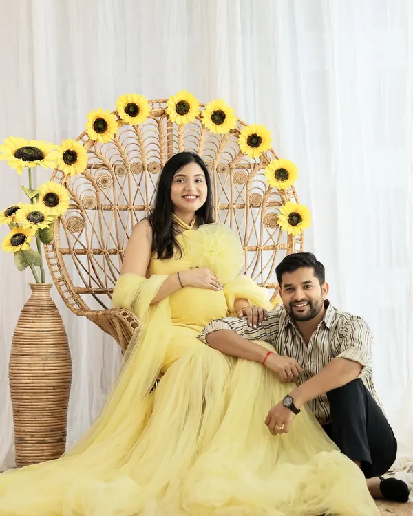 A bright and joyful couple portrait. The mom-to-be is radiant in a yellow tulle gown, seated on a peacock chair decorated with sunflowers, while her partner sits lovingly by her side.