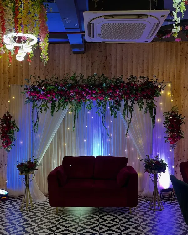 A classic Roka ceremony stage featuring a plush red sofa set against a backdrop of white drapes and fairy lights. The overhead floral arrangement with deep red flowers and greenery adds a touch of luxury.