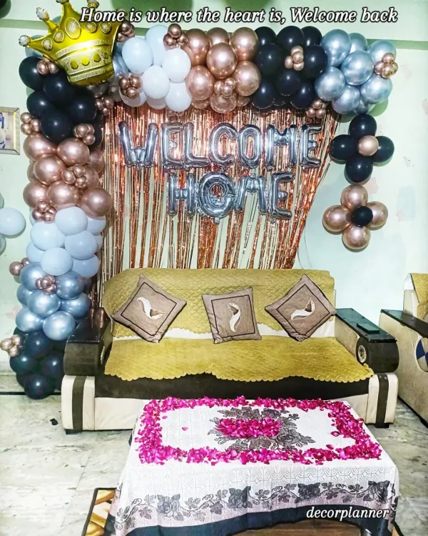 A stylish 'Welcome Home' decoration in black, silver, and rose gold. The balloon arch over the sofa, combined with a shimmery curtain and flower petal art on the table, creates a chic and celebratory look.
