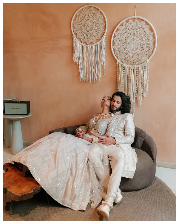 A stunning indoor portrait with the couple dressed in elegant white traditional wear. The bohemian decor and warm lighting create a sophisticated and romantic atmosphere.