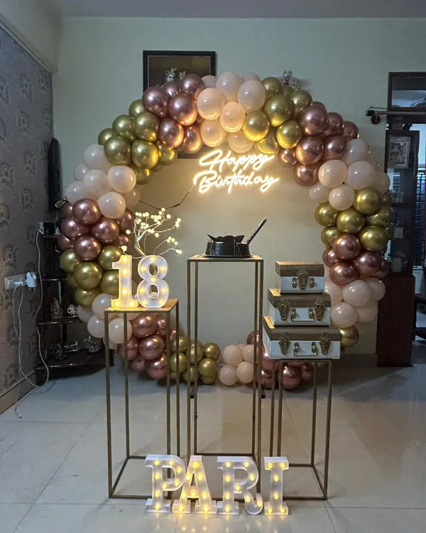 An elegant 18th birthday celebration with a rose gold, gold, and pastel pink balloon arch. The setup includes tall plinths, marquee numbers, and a neon sign.