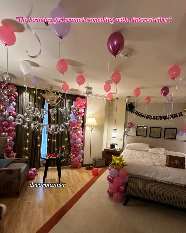A full view of this Pinterest-worthy birthday setup. The room is filled with pink, purple, and silver balloons, with two beautiful balloon columns framing the "Happy Birthday" banner and fairy lights.