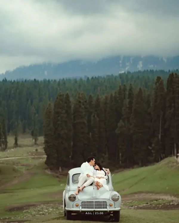 A breathtaking shot of a couple sharing an intimate moment on a vintage car, surrounded by the vast, pine-covered hills of Kashmir. This is the essence of a destination pre-wedding shoot, combining romance with awe-inspiring scenery.