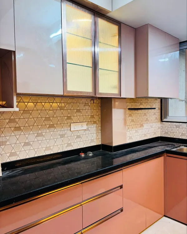 A modern L-shaped kitchen with a beautiful dual-tone design. The lower cabinets are a matte salmon pink with gold-accented handles, while the upper cabinets mix solid panels with fluted glass doors and a geometric backsplash.