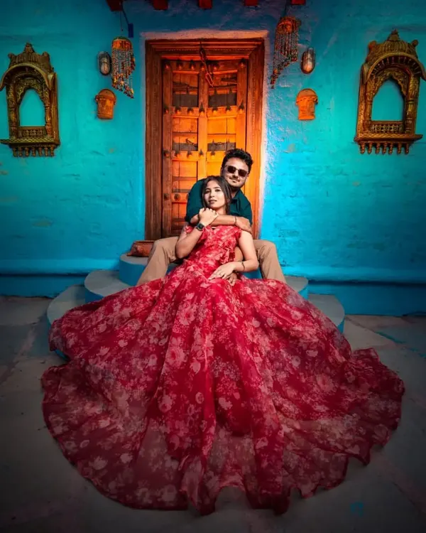 A vibrant and artistic pre-wedding shot. The red floral printed organza gown looks stunning against the colorful blue wall, creating a photo that is full of life and personality.
