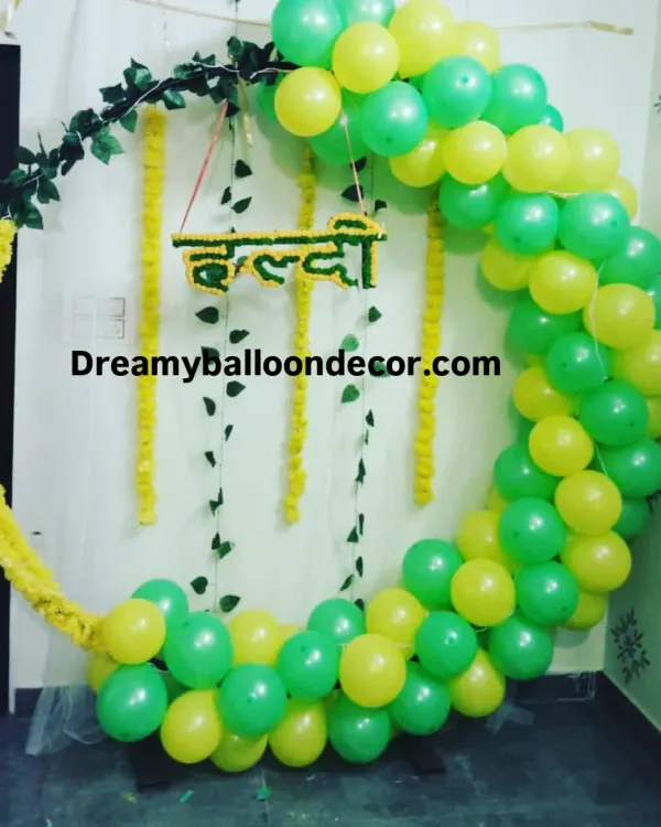 A simple yet beautiful Haldi decoration for a home ceremony. The circular balloon arch in green and yellow is accented with hanging marigold garlands and a 'Haldi' sign, creating a perfect photo spot.