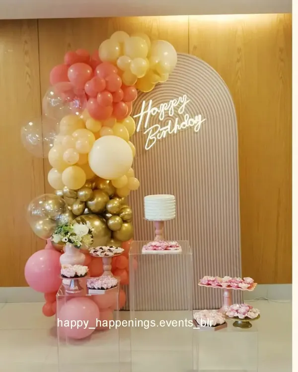 This minimalist birthday setup features a soft pink ribbed arch panel with a delicate neon sign. The organic balloon garland in pastel pink, peach, and gold adds a touch of color without overwhelming the clean, modern aesthetic.
