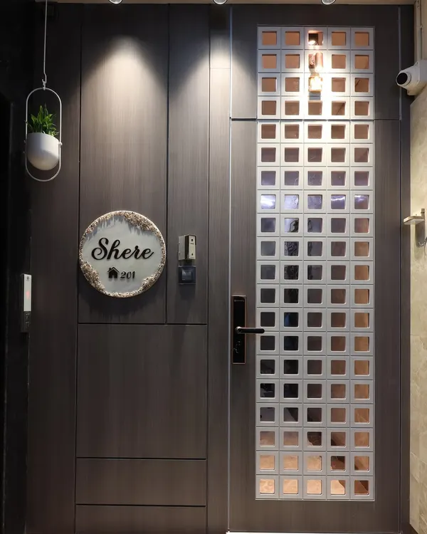 A modern entrance featuring a dark wood-finish main door and a contrasting white safety door with a square grid pattern. The design is complemented by a custom nameplate and focused downlights for a warm welcome.