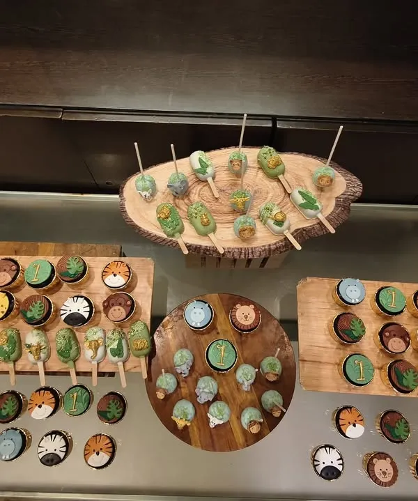 Here is an overhead view of a complete jungle-themed dessert table. You can see the variety of treats, from cupcakes with animal faces to leaf-adorned cakesicles, all arranged on rustic wooden platters to enhance the safari vibe.