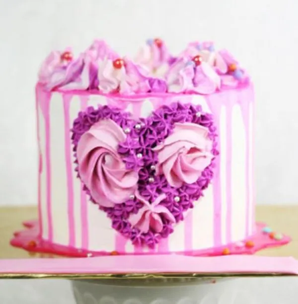 This modern and artistic drip cake is designed for love. It features a tall profile with pink drips over white frosting and a beautiful heart made of purple and pink buttercream rosettes.