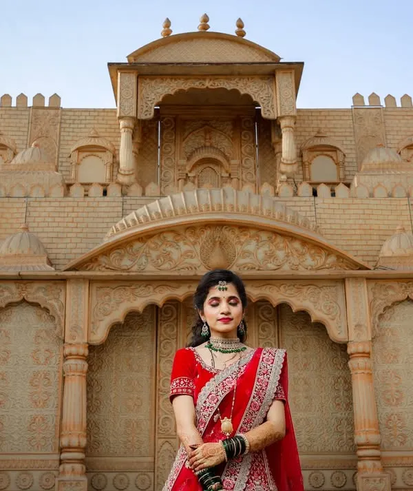 A stunning wide shot of a bride in a red lehenga against a grand, palace-like backdrop. This image showcases the bridal attire in a majestic setting, perfect for a destination wedding lookbook.