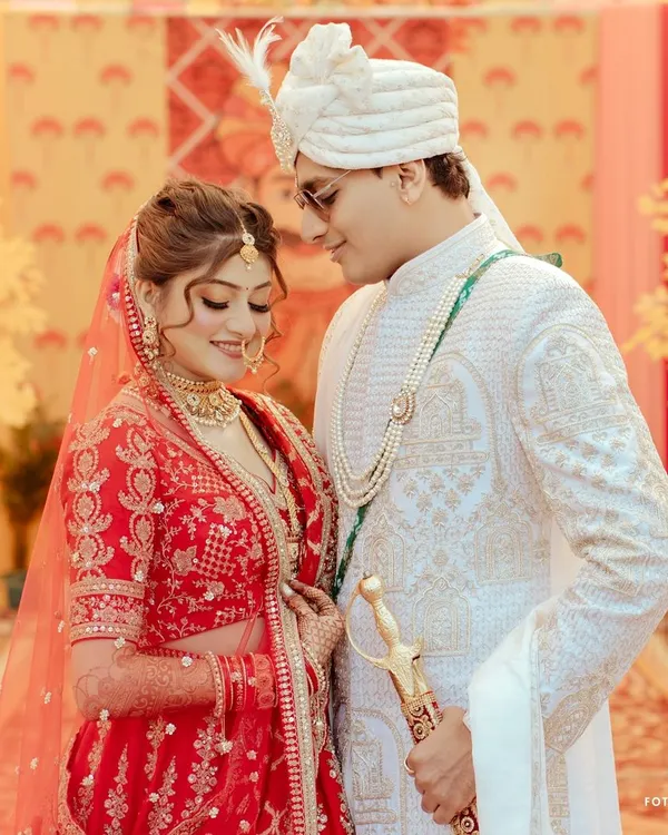 A classic portrait of the couple in their wedding attire, the bride in a traditional red lehenga and the groom in a white sherwani, capturing their regal presence.