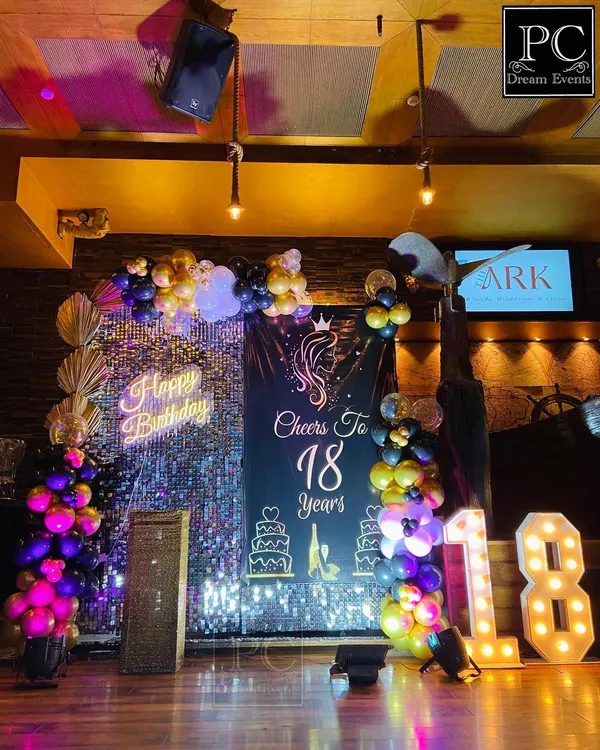 A glamorous "Cheers to 18 Years" birthday party setup. This decor features a silver sequin wall, a custom backdrop, a 'Happy Birthday' neon sign, and light-up marquee numbers, perfect for a milestone celebration.